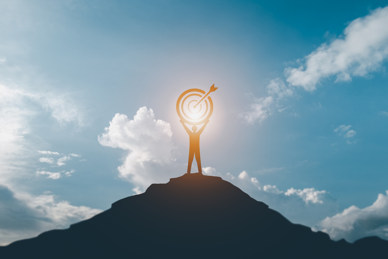 The silhouette of a businessman holding a target board on a mountain of blue and sunlight is a symbol of success in leading with purpose.